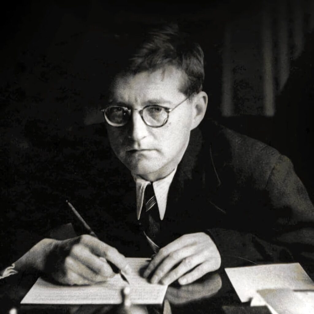 Composer Dmitri Shostakovich sitting at a desk, pen in hand, looking pensive.