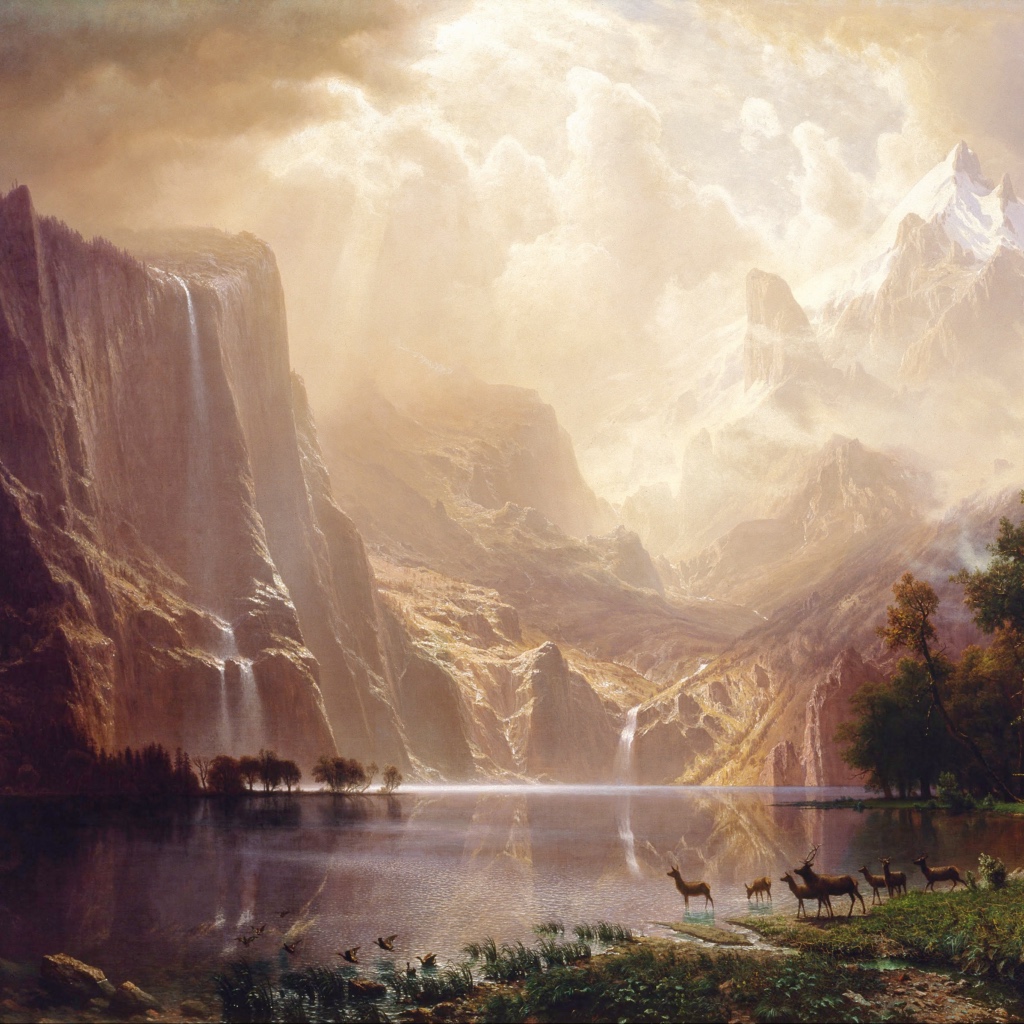 The sun peeks through heavy clouds to illuminate tall mountains; waterfalls plunge down the steep sides into a lake at which a herd of deer has stopped to drink