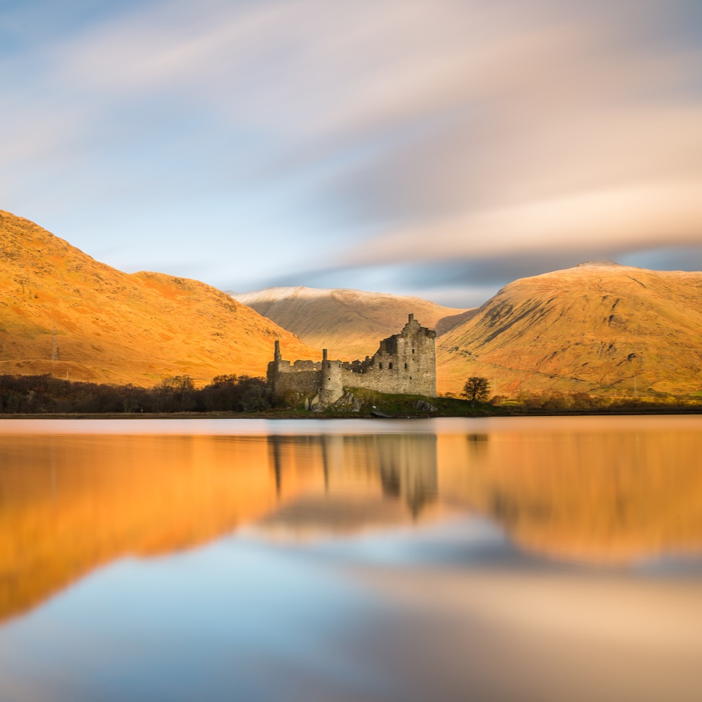 A ruined Scottish castle is reflected on the loch, with golden hills behind