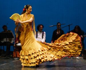 Photo of flamenco dancer Eva Conti on stage in a long golden yellow dress holding a yellow fan