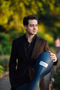 Portrait of cellist John-Henry Crawford standing in a park holding his cello case. Photo by Jiyang Chen